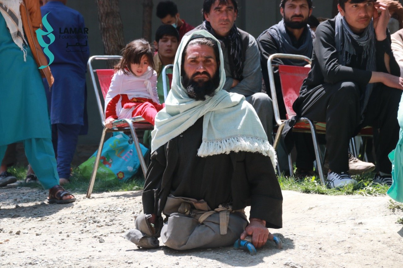 A man with amputated legs sits on the ground, wearing a shawl and black clothes. Behind him, several people, including a young girl in a pink dress, sit on red chairs outside, watching attentively. A man with amputated legs sits on the ground, wearing a shawl and black clothes. Behind him, several people, including a young girl in a pink dress, sit on red chairs outside, watching attentively.