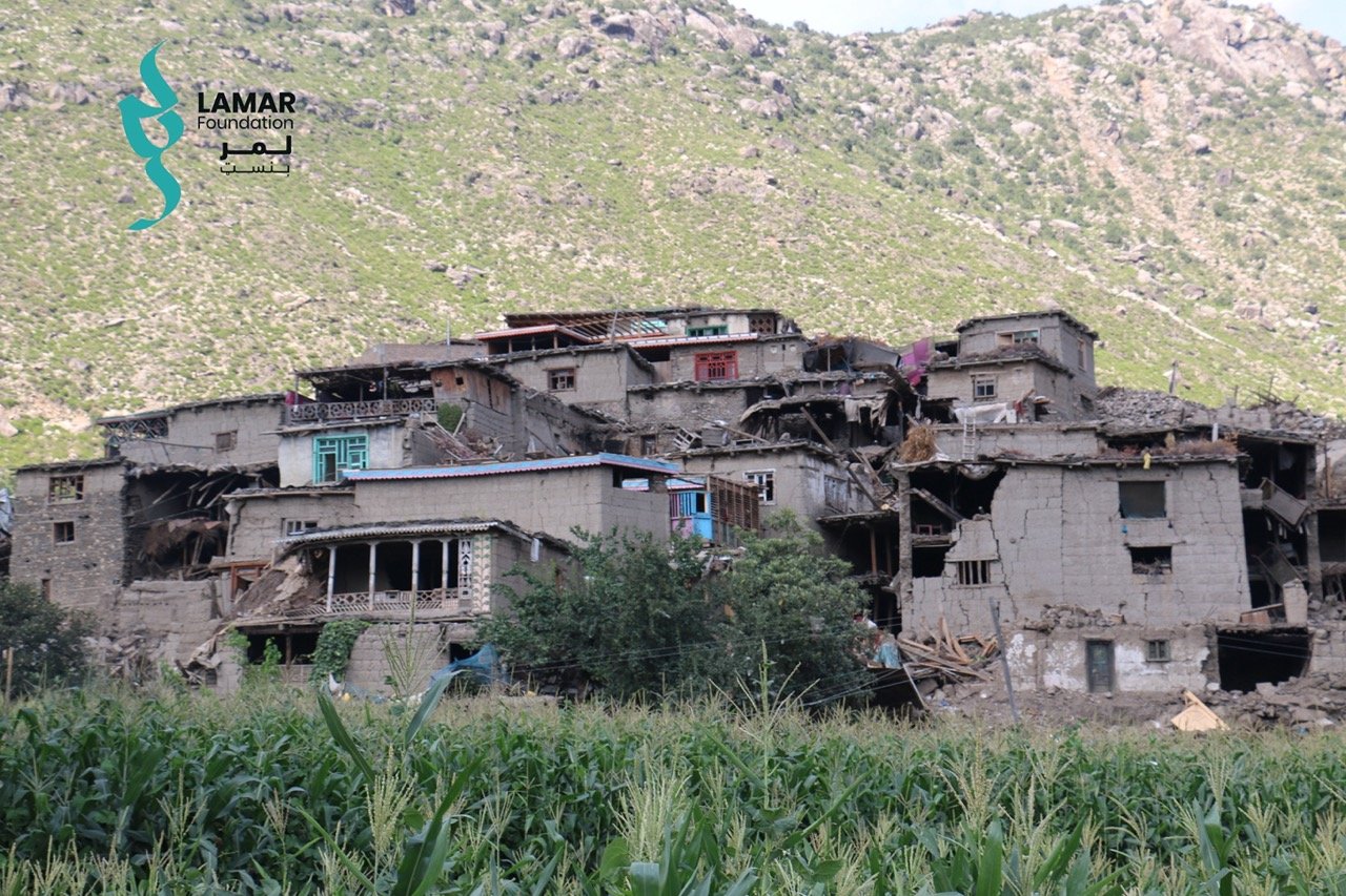 A cluster of damaged, mud-brick houses sits at the base of a green, tree-covered hillside. The foreground has tall crops and a bush. The Lamar Foundation logo appears in the top left corner. A cluster of damaged, mud-brick houses sits at the base of a green, tree-covered hillside. The foreground has tall crops and a bush. The Lamar Foundation logo appears in the top left corner.