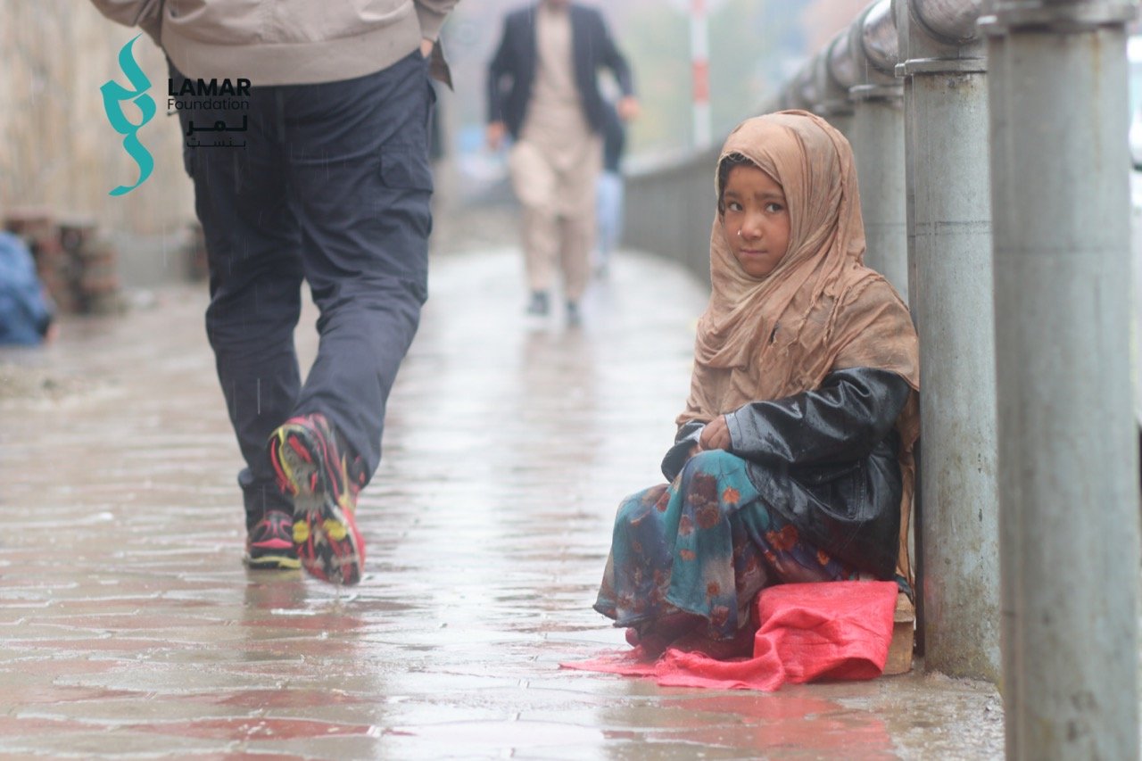 A young girl in a headscarf and colorful dress sits on a wet sidewalk in the rain, looking at the camera, while people walk past her. A red cloth is on the ground beside her. A young girl in a headscarf and colorful dress sits on a wet sidewalk in the rain, looking at the camera, while people walk past her. A red cloth is on the ground beside her.