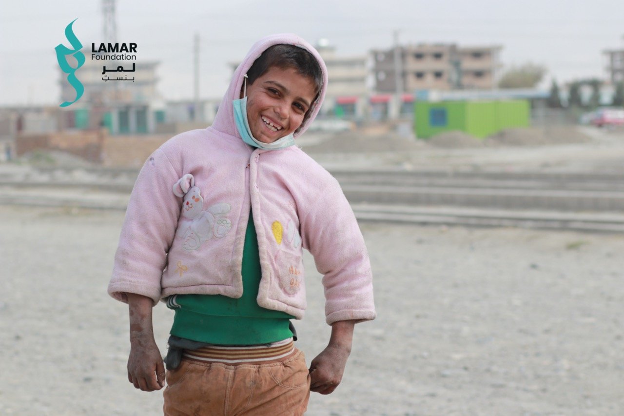 A smiling child wearing a pink jacket and green shirt stands in a dusty outdoor area, with visible dirt on their hands and face. Buildings and construction materials can be seen in the blurred background. The Lamar Foundation logo is on the image. A smiling child wearing a pink jacket and green shirt stands in a dusty outdoor area, with visible dirt on their hands and face. Buildings and construction materials can be seen in the blurred background. The Lamar Foundation logo is on the image.