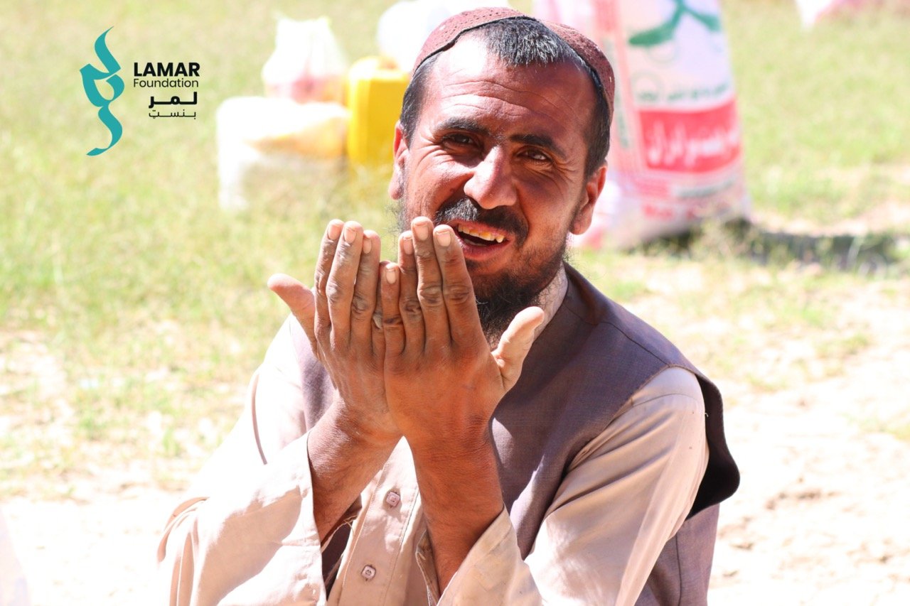 A man in traditional clothing sits outdoors, smiling and holding his hands up in front of him. The logo of Lamar Foundation appears in the top left corner. There are supplies and grass visible in the background. A man in traditional clothing sits outdoors, smiling and holding his hands up in front of him. The logo of Lamar Foundation appears in the top left corner. There are supplies and grass visible in the background.