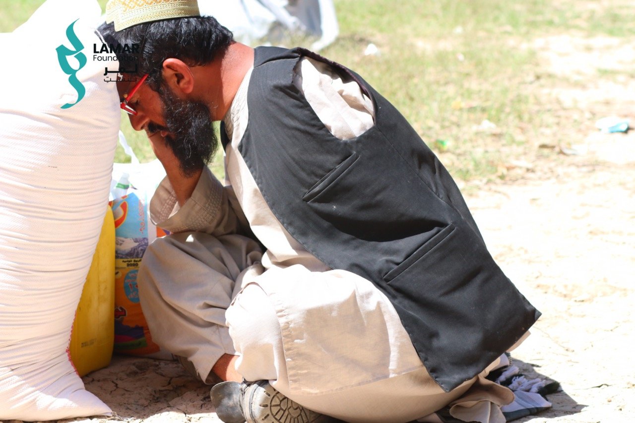 A man in traditional clothing and a cap crouches on the ground outdoors, leaning on a sack and appearing deep in thought or distress. Sunlight highlights the scene, and a charity logo is visible in the background. A man in traditional clothing and a cap crouches on the ground outdoors, leaning on a sack and appearing deep in thought or distress. Sunlight highlights the scene, and a charity logo is visible in the background.