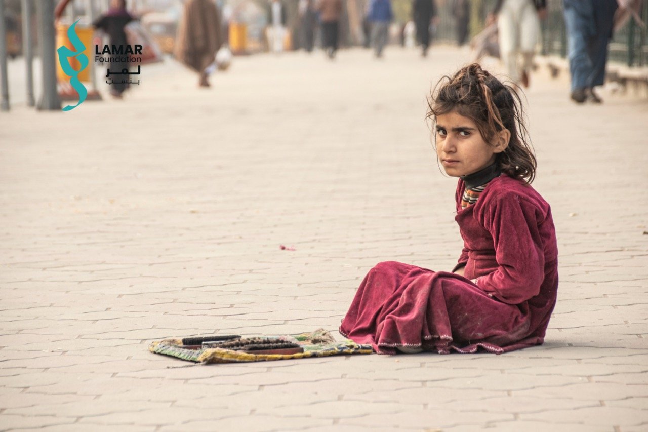 A young girl in a maroon dress sits on a paved sidewalk with some items laid out in front of her, looking toward the camera. People are visible in the blurred background. The Lamar Foundation logo appears in the top left corner.