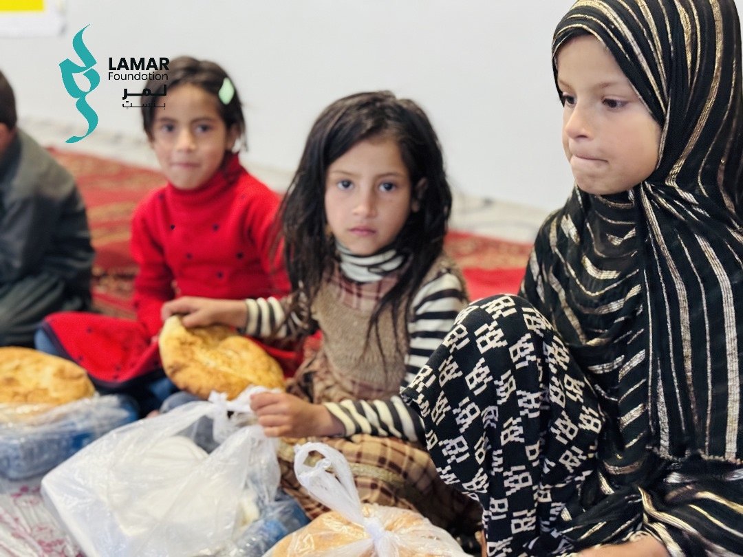Three young girls sit on a patterned carpet, each holding bread and food packages. One girl wears a striped hijab, and the others are dressed in red and brown. The LAMAR Foundation logo appears in the background. Three young girls sit on a patterned carpet, each holding bread and food packages. One girl wears a striped hijab, and the others are dressed in red and brown. The LAMAR Foundation logo appears in the background.
