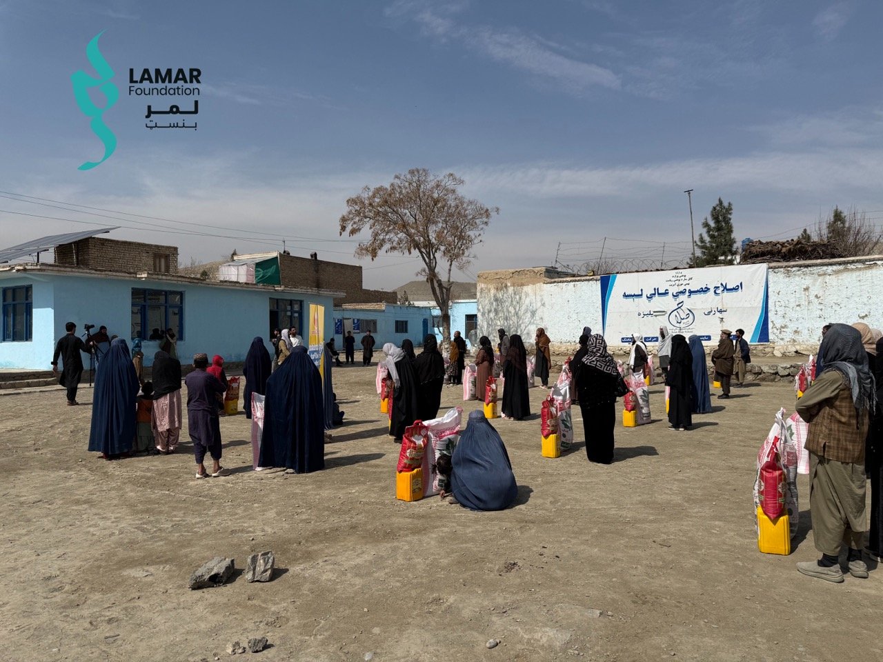 People, many wearing traditional clothing, stand in line outdoors with bags and yellow containers, waiting for aid distribution near blue buildings. A sign and the Lamar Foundation logo are visible in the background. People, many wearing traditional clothing, stand in line outdoors with bags and yellow containers, waiting for aid distribution near blue buildings. A sign and the Lamar Foundation logo are visible in the background.