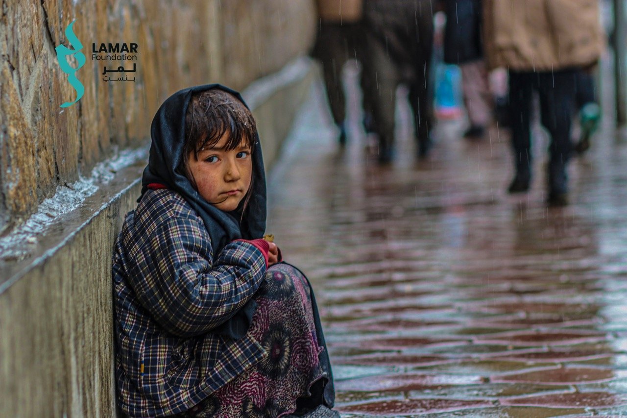 A young child in a plaid coat and dark headscarf sits alone on a wet sidewalk near a wall, looking sad. Rain falls, and people walk by in the blurred background. The Lamar Foundation logo appears in the corner. A young child in a plaid coat and dark headscarf sits alone on a wet sidewalk near a wall, looking sad. Rain falls, and people walk by in the blurred background. The Lamar Foundation logo appears in the corner.