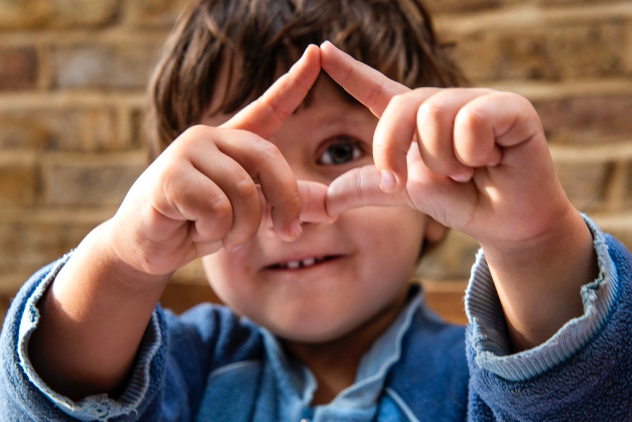 A young child in a blue sweater smiles while holding their hands up to form a triangle shape with their fingers. The background features a blurred brick wall.