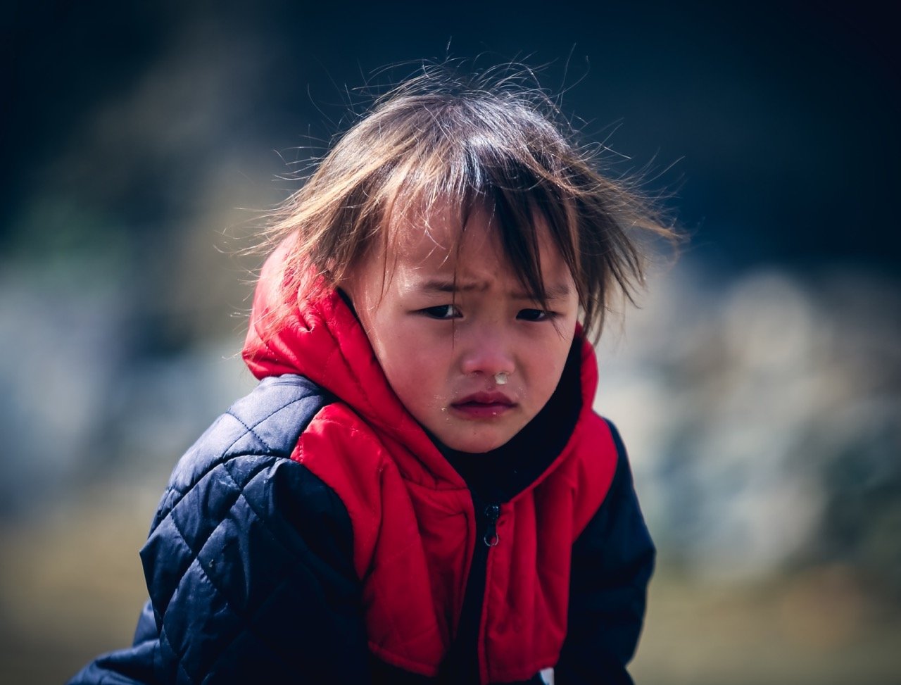 A young child with straight brown hair and a red and navy blue quilted jacket looks upset or sad, with teary eyes and a frown, standing outdoors against a blurred natural background.