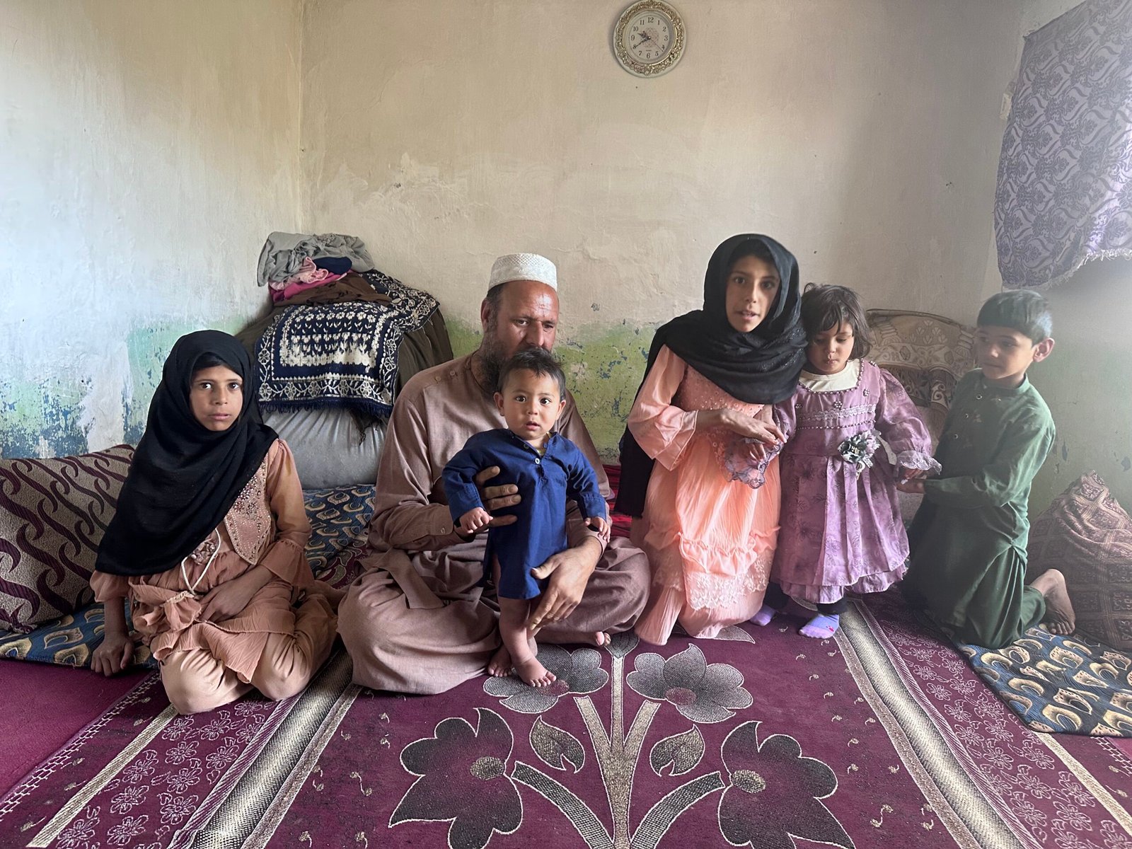 A man sits cross-legged on a patterned carpet in a simple room, holding a small child. Four other children, two girls and two boys, sit or stand around him, all dressed in traditional clothing. A clock hangs on the wall behind them.